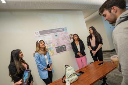 Students stand next to a poster with a backpack on a table in front of them